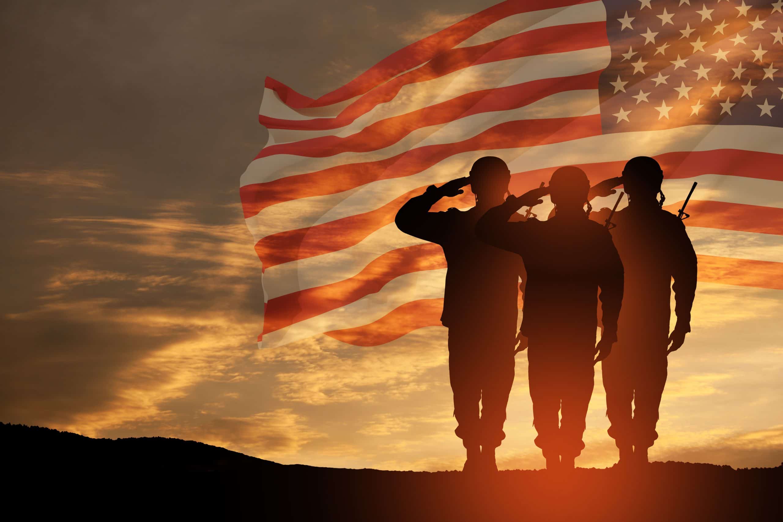 Three silhouetted soldiers stand and salute in front of a large, translucent American flag at sunset, with the sky glowing in orange and gold tones.