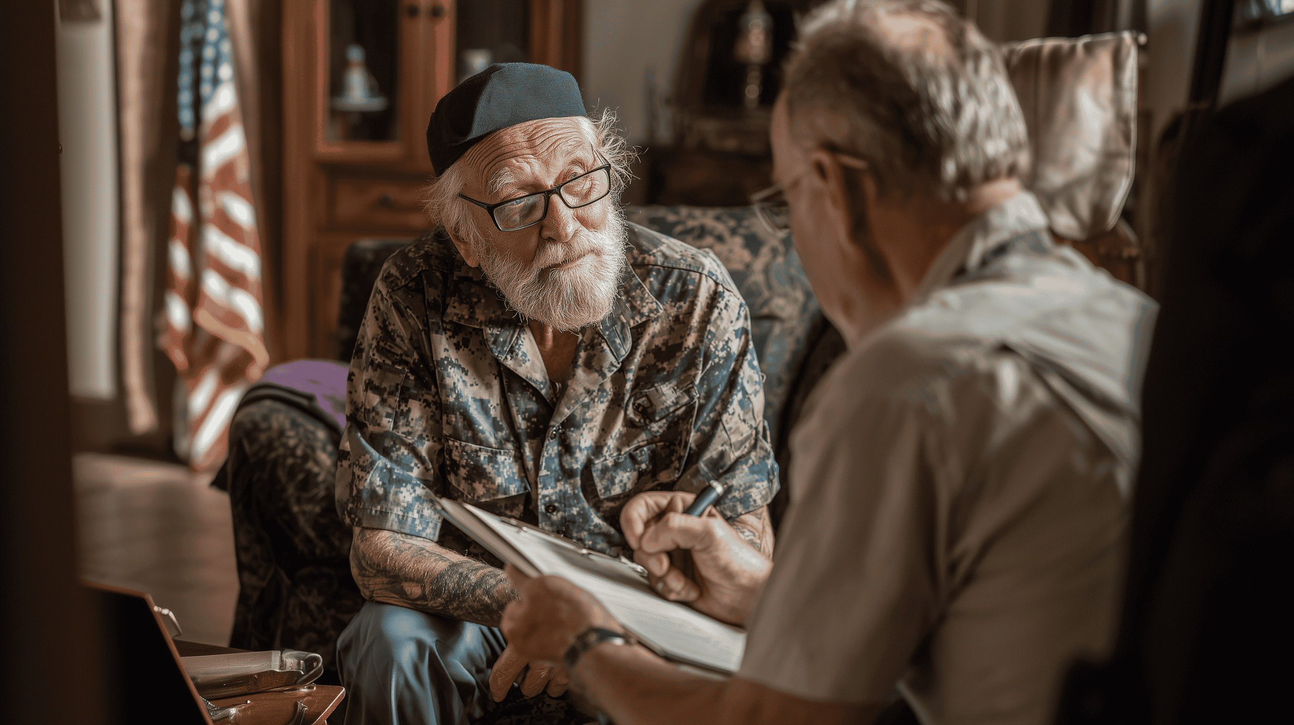 An elderly man with a white beard, wearing glasses and a blue cap, sits on a chair while another older man interviews him with a clipboard. An American flag is visible in the background, suggesting a veteran’s setting.