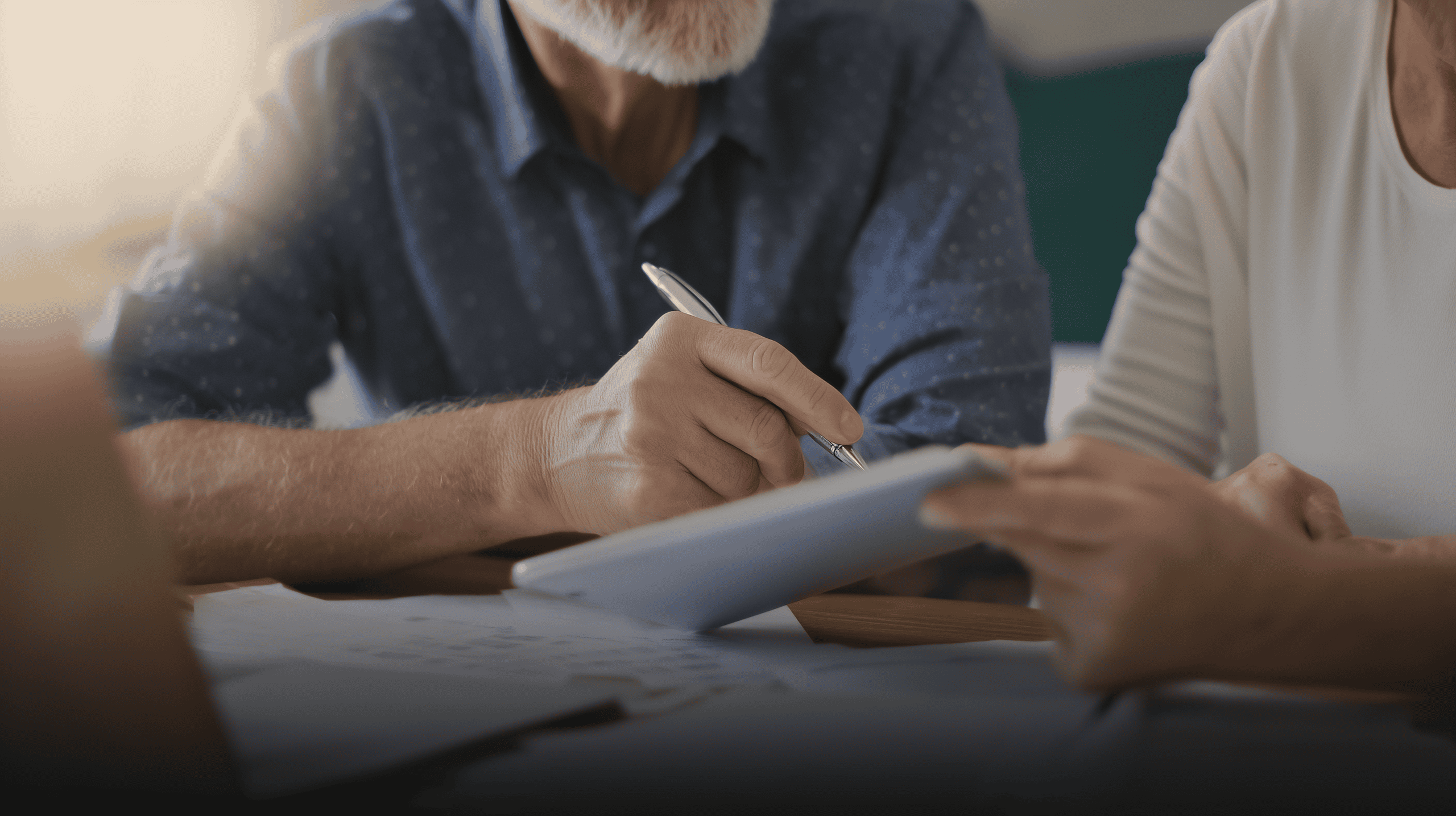 Two older adults sit at a table discussing documents; one is holding a pen and writing on a tablet while the other points at the screen. Papers and a laptop are visible on the table.