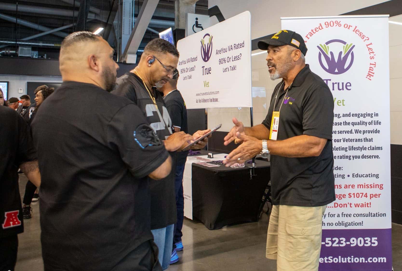 Three men talk at an indoor event near a booth with a “True Vet” banner featuring purple hands and text about veterans’ benefits. One man gestures while the others listen and one checks a phone.