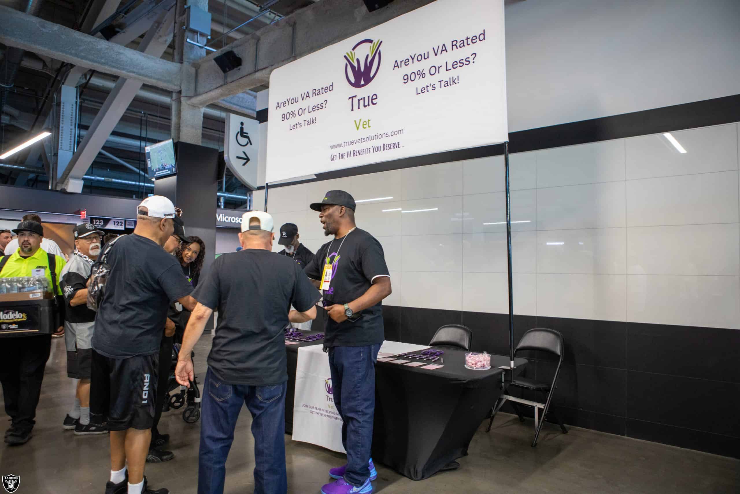 People gather and interact at an indoor event booth for True Vet, which offers resources for veterans rated 90% or less by the VA. The booth has a purple and white banner and informational materials on a table.