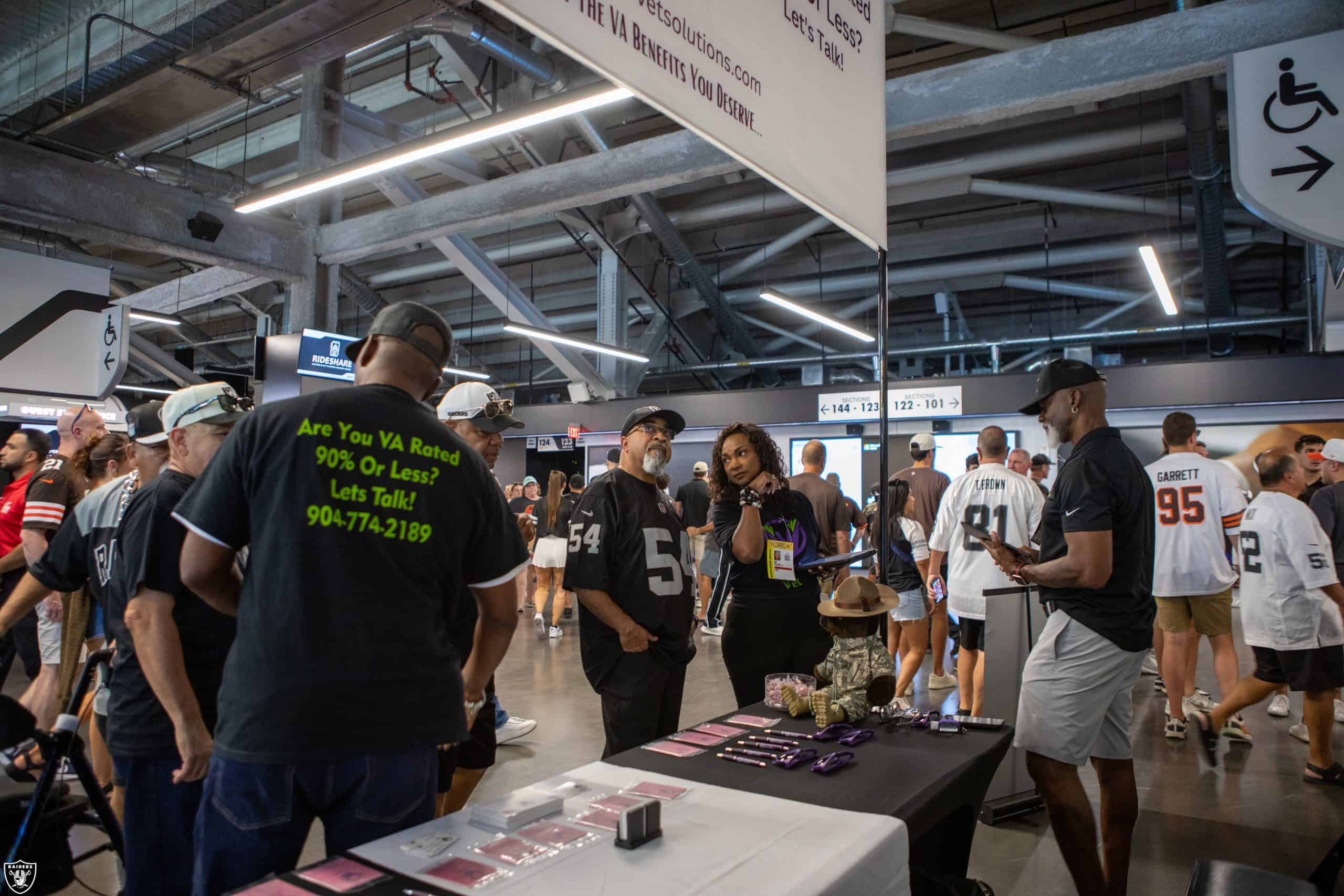 Fans in Raiders jerseys gather around an informational booth inside a stadium concourse. Some people browse items on the table, while others talk and walk by. The area is busy with signs and open space above.